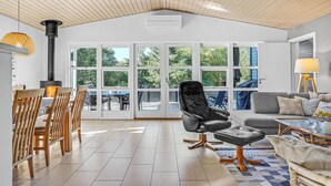 Ein geräumiger Raum mit hoher Decke, großen Fenstern, einem Essbereich mit Holzsesseln, einem schwarzen Ledersessel und einer Couch mit Kissen.