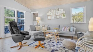 Ein modernes Wohnzimmer mit einem grauen Sofa, einem schwarzen Ledersessel, einem Couchtisch und einem Bücherregal. An den Wänden befinden sich gerahmte botanische Drucke und ein Fenster mit Blick auf Bäume.