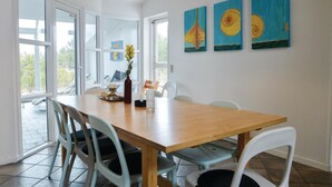 A dining area with a wooden table, white chairs, and a vase with flowers. There are three paintings on the wall.