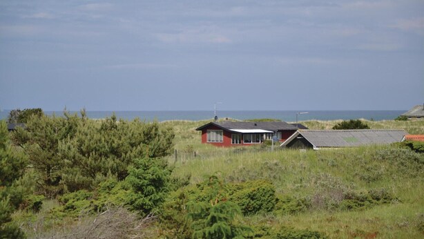 A small, single-story building with a sloped roof, surrounded by dense greenery and a view of the sea.