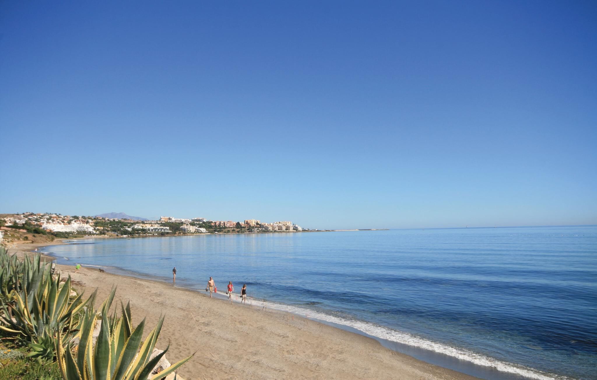 Une plage avec une eau d’un bleu limpide, un rivage sablonneux et des bâtiments en arrière-plan.