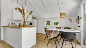 Une cuisine moderne avec des meubles blancs, un plan de travail en bois et un îlot central. On y trouve un coin repas avec une table blanche, des chaises noires et un plaid en fausse fourrure.