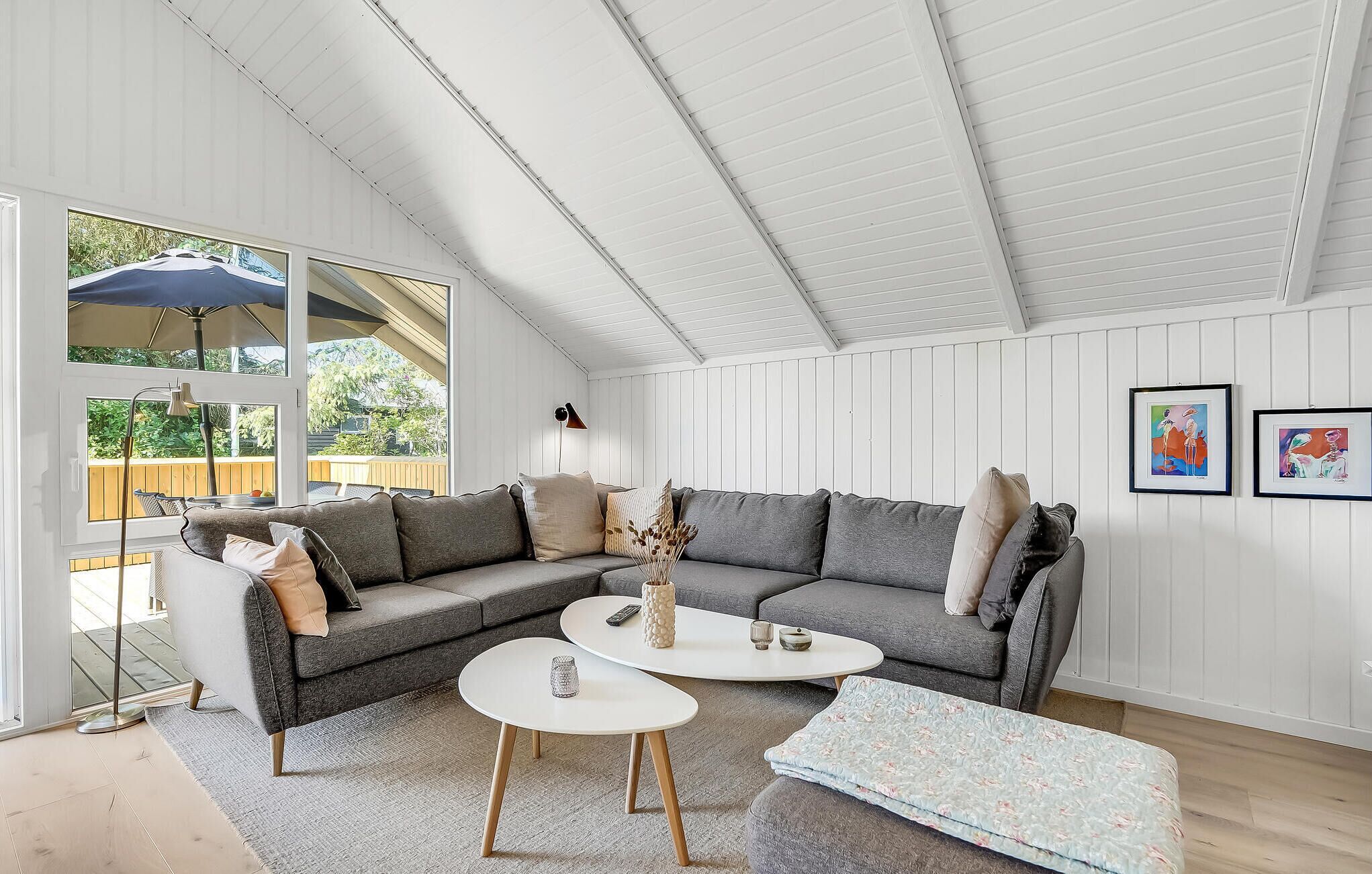 Ein modernes Wohnzimmer mit einem grauen Ecksofa, zwei weißen Couchtischen und einem großen Fenster mit Blick auf eine Terrasse.