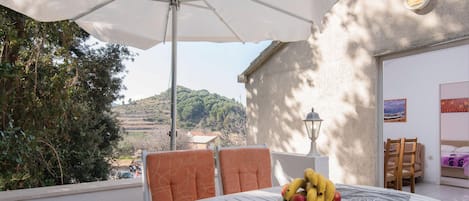 A round table with a fruit bowl, two orange chairs, a white umbrella, and a view of a hillside.