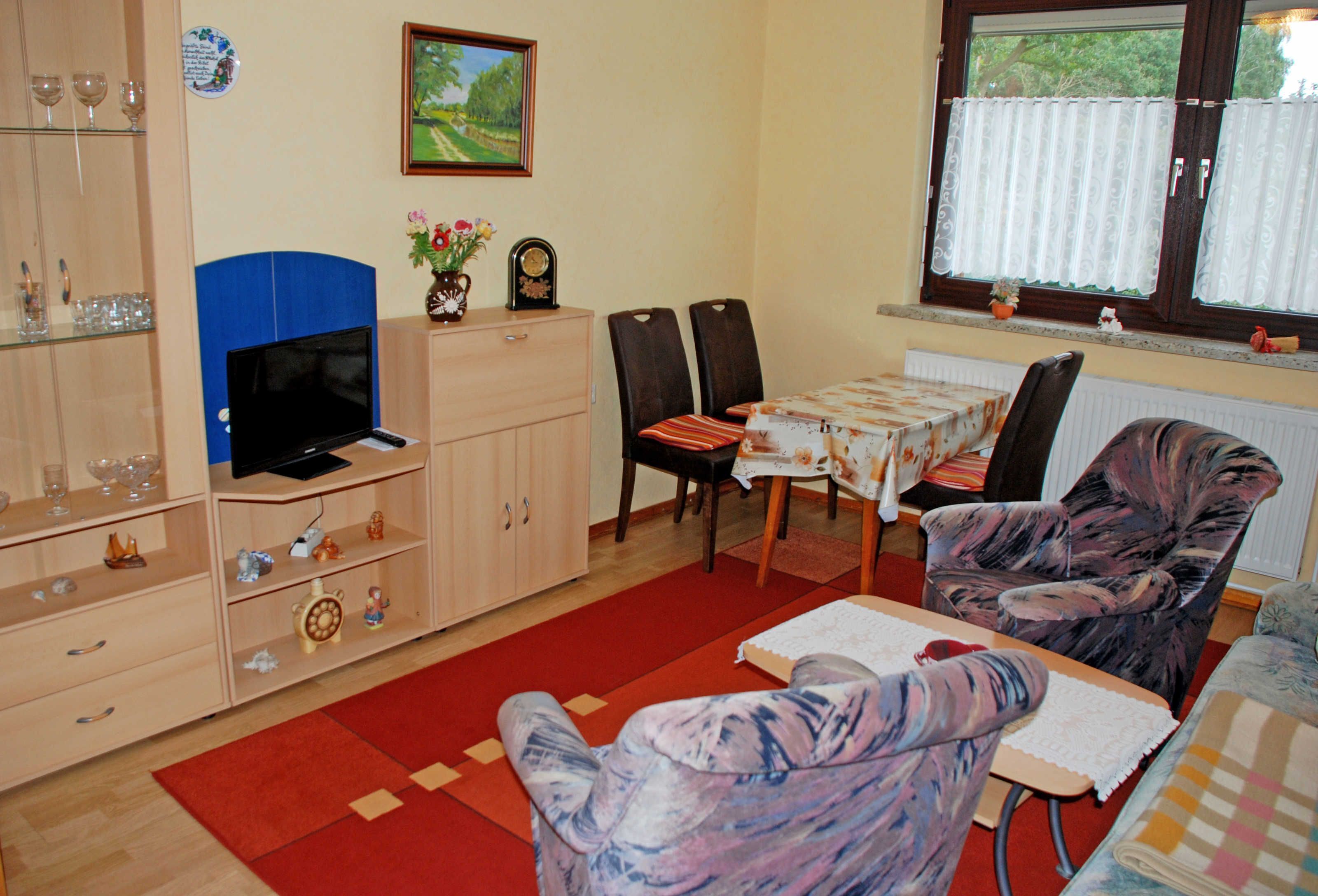 Uma sala de estar com televisão, armários de madeira, uma mesa de jantar com cadeiras e uma janela com cortinas.