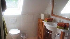 Ein Badezimmer mit Toilette, Waschbecken und Dusche. Ein Fenster sorgt für natürliches Licht, und Handtücher sind ordentlich auf dem Waschbecken gestapelt.