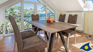 A dining area with a wooden table and brown upholstered chairs, a fruit bowl on the table, and a view of the sea through large windows. - Villa Strandperle, Whg. 33 // - VS 33 (Heringsdorf)