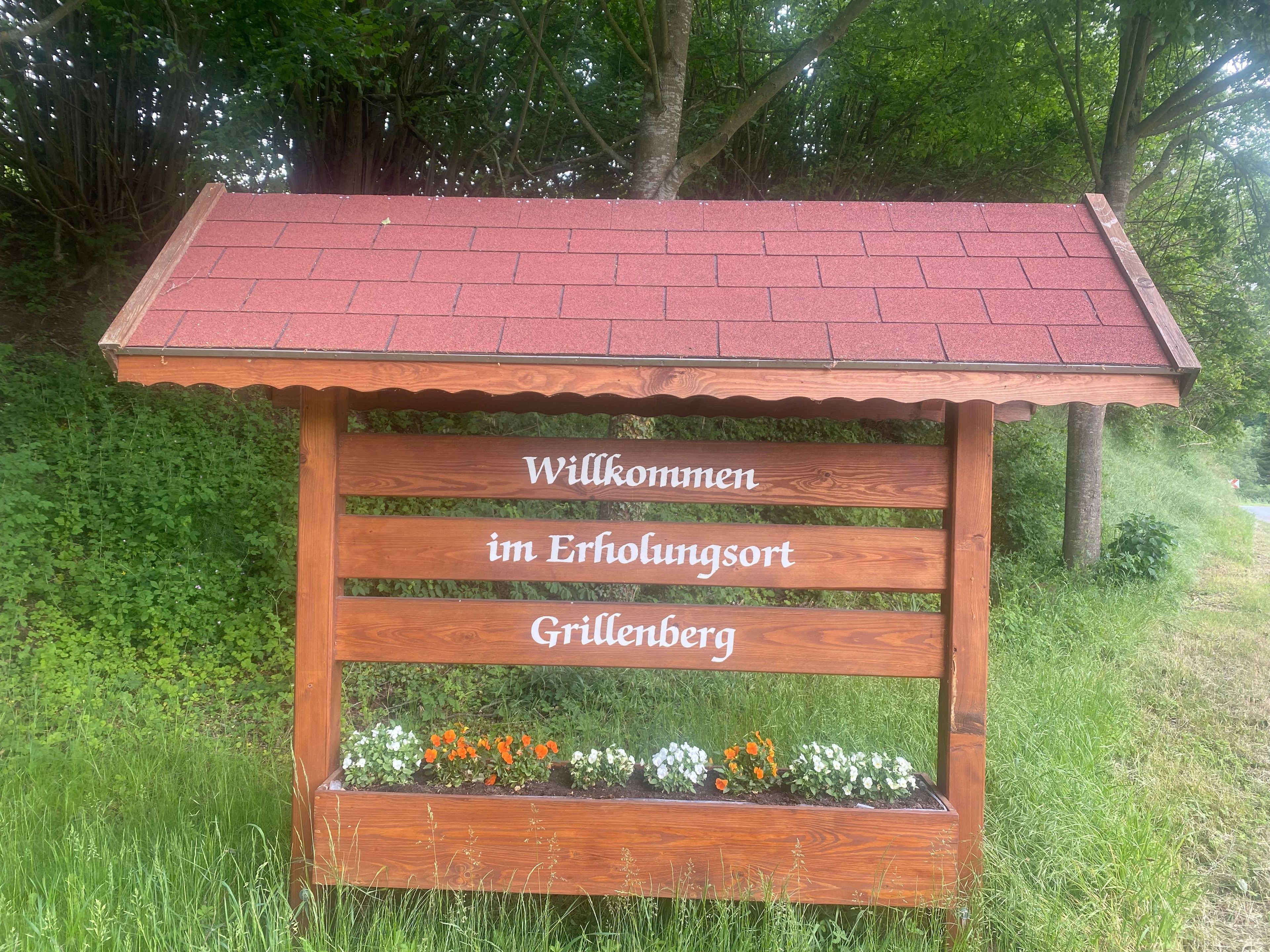 A wooden welcome sign with 'Willkommen im Erholungsort Grillenberg' text, surrounded by a garden with flowers and grass.
