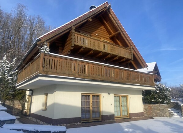 A two-story log cabin with a balcony, wooden exterior, and a stone foundation.