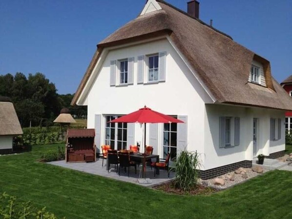 A white house with a thatched roof, a patio with outdoor furniture, and a red umbrella.