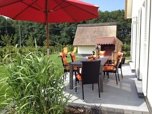 An outdoor patio with a table and chairs, a red umbrella, and a thatched-roof structure.