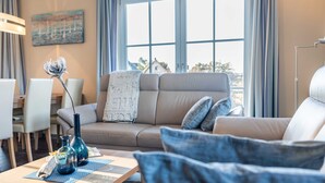 A modern living room with a beige sofa, a dining table with chairs, and a large window with curtains. - Holiday home Strandmuschel - FH Strandmuschel (Loddin)
