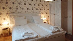 Een opgemaakt bed met witte lakens, een bedlampje en behang met een bloemenmotief.