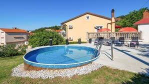 A swimming pool with a wooden deck, surrounded by a pebble border, in front of a yellow building with a red roof. - Cozy apartment in Viskovo with WiFi (Viskovo)