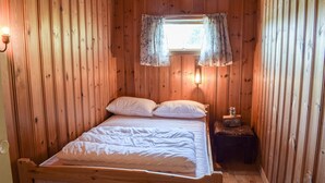 Ein Zimmer mit Holzwänden, einem Bett, einem kleinen Tisch mit einem Radio und einem Fenster mit floralem Vorhang.