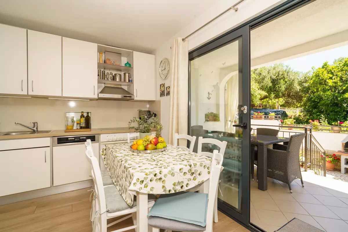 Een keuken met witte kastjes, een eettafel met een schaal fruit en een glazen schuifpui naar een terras buiten met meubels.