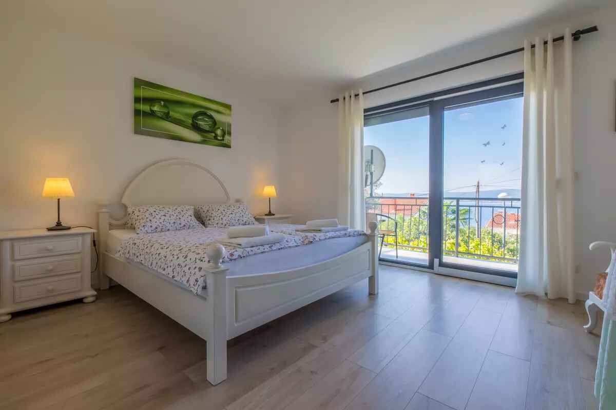 A bedroom with a bed, bedside tables, a large window with a view of greenery, and a balcony.