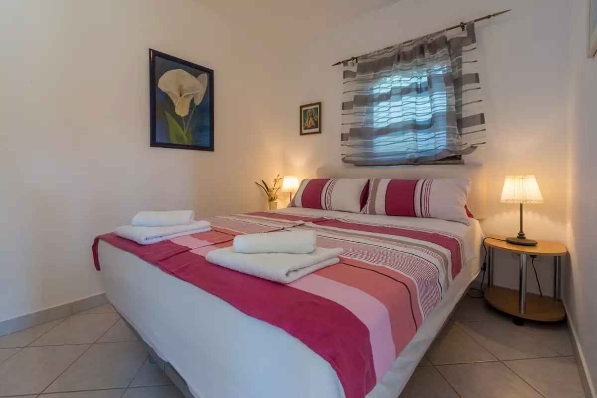 A neatly made bed with folded towels, a painting of a flower, a small religious icon, and a window with striped curtains.