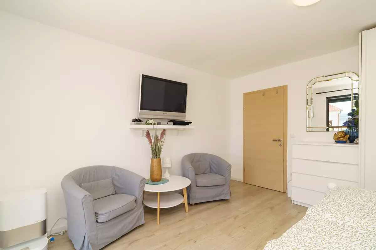 Ein kleiner, moderner Raum mit einem an der Wand befestigten Flachbildfernseher, zwei grauen Sesseln, einem kleinen runden Tisch mit einer Vase und einer Holztür.