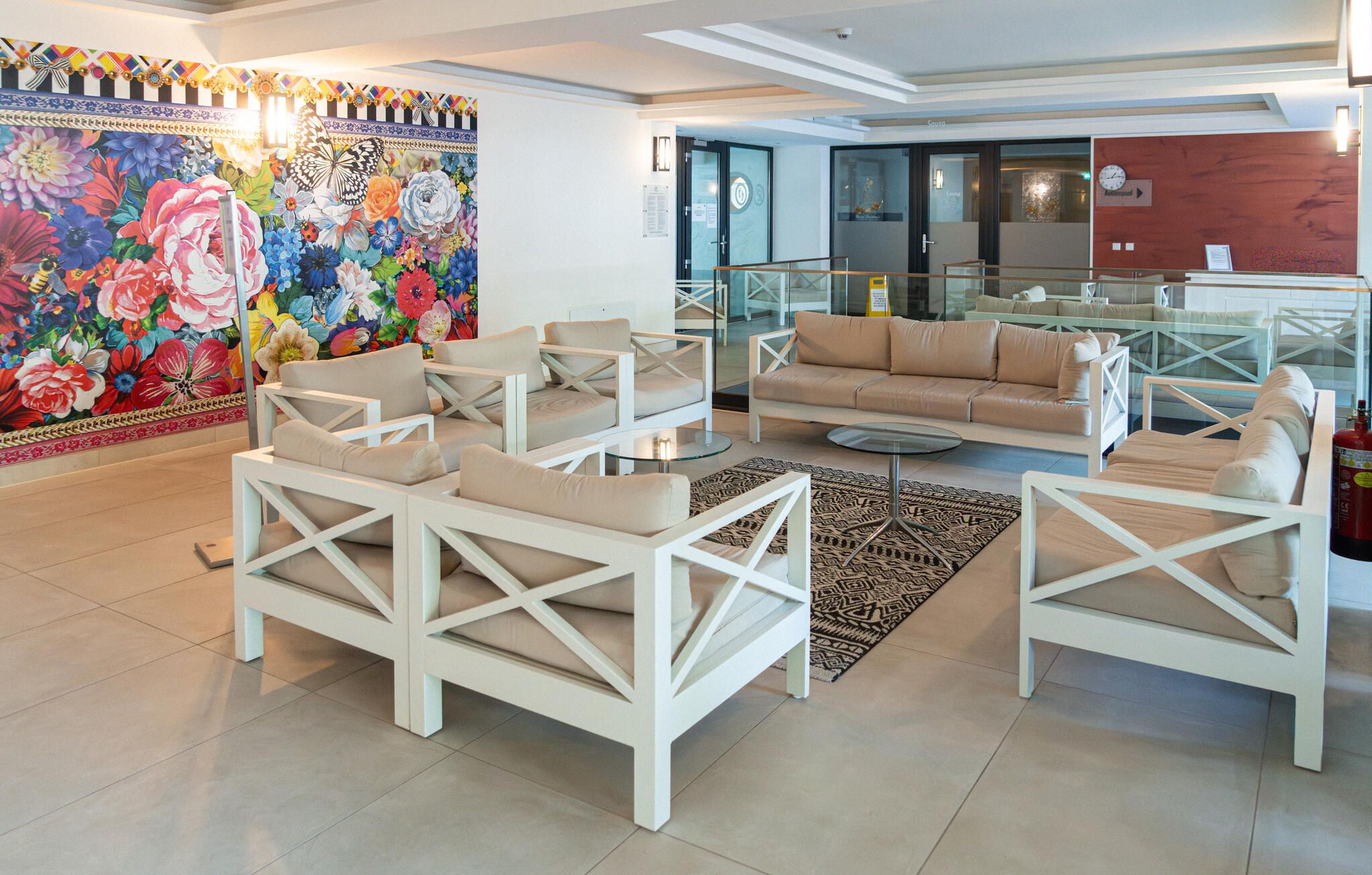 A modern living room with white furniture, a colorful floral mural, and a glass partition separating the living area from a kitchen.