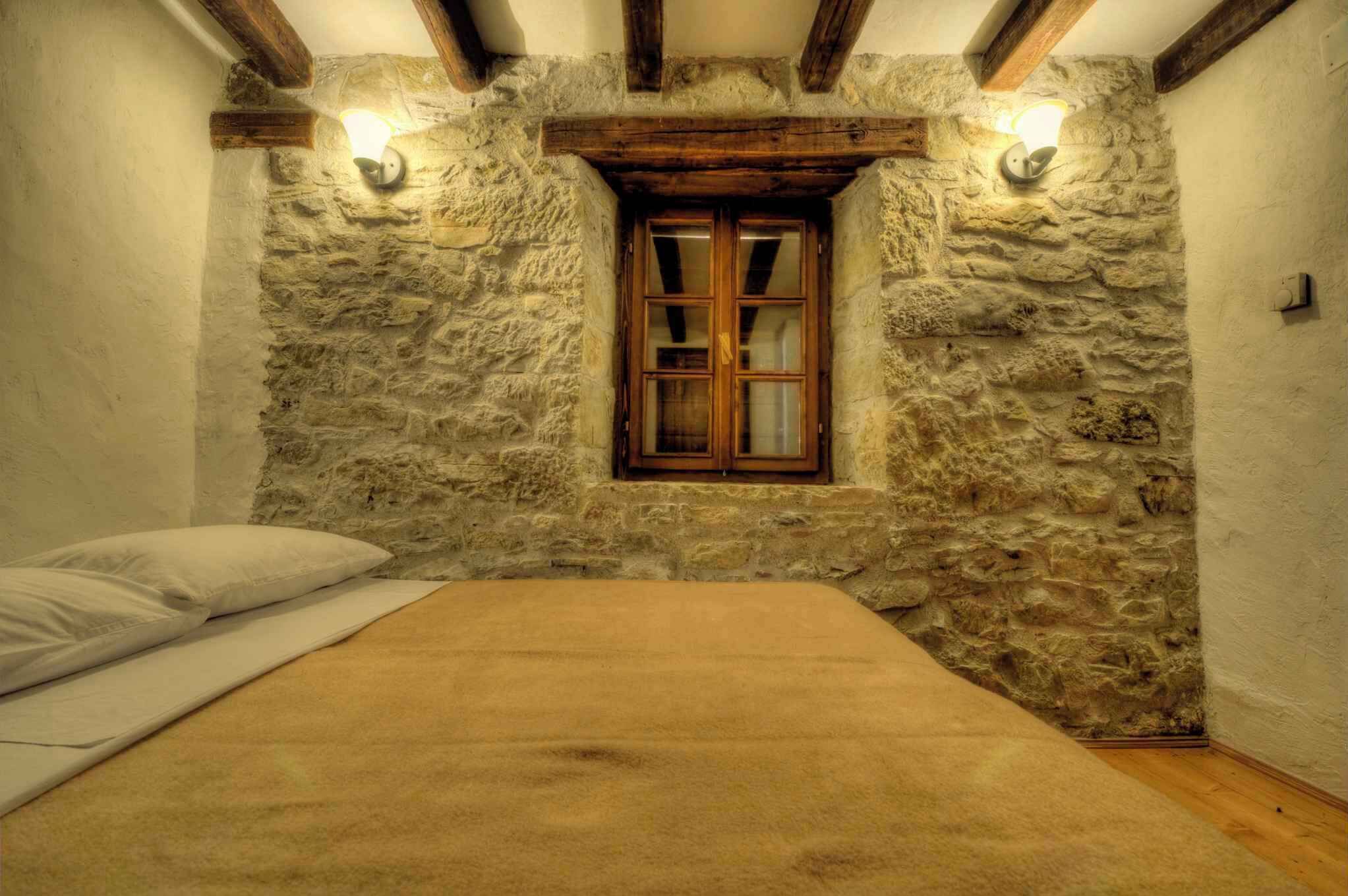 A room with a stone wall, a wooden window, a bed, and a wall-mounted light fixture.