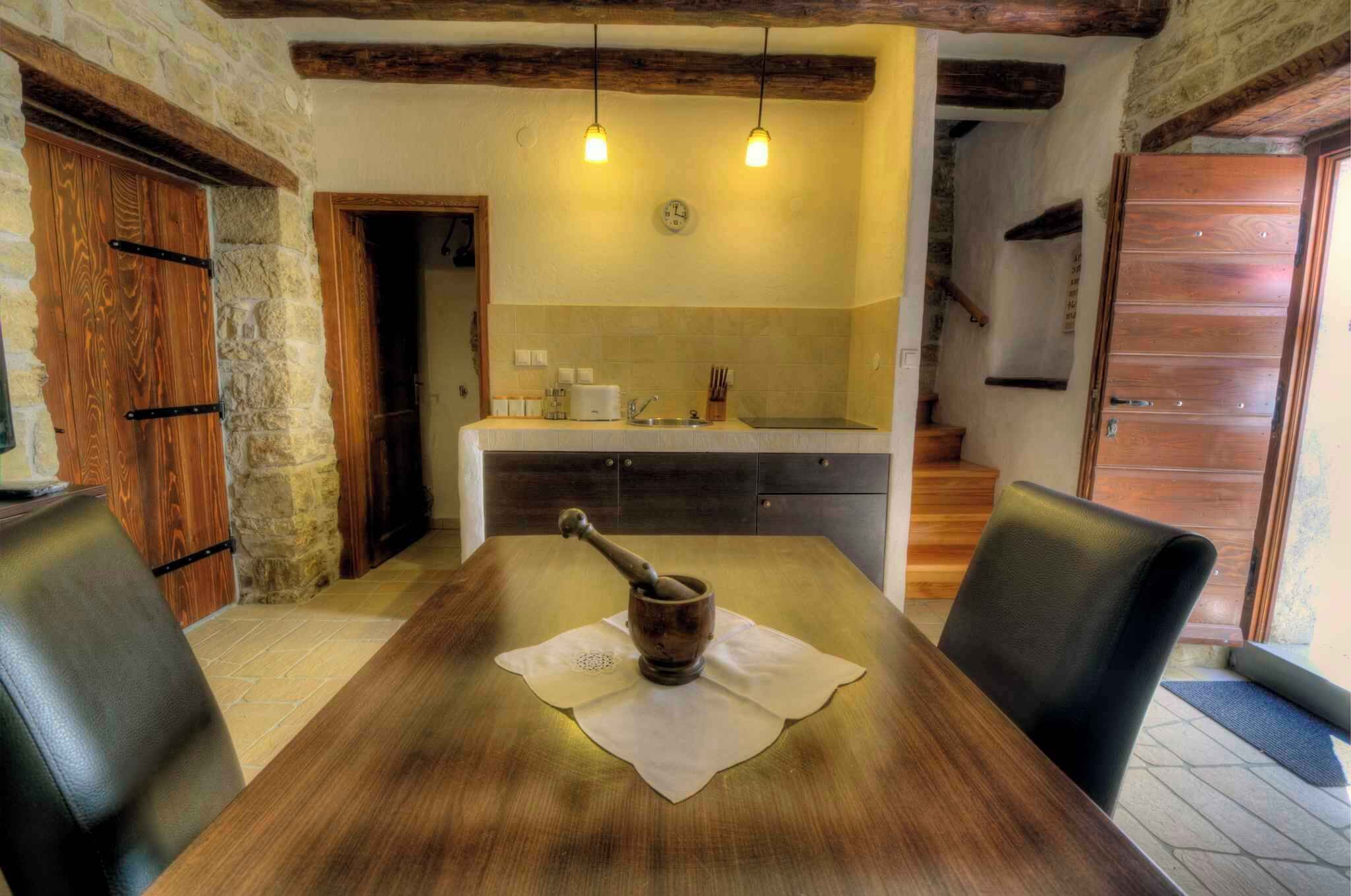 A rustic dining area with a wooden table, two chairs, a rustic mortar and pestle, and a kitchen in the background.
