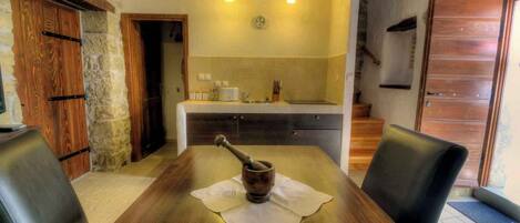 A rustic dining area with a wooden table, two chairs, a rustic mortar and pestle, and a kitchen in the background.