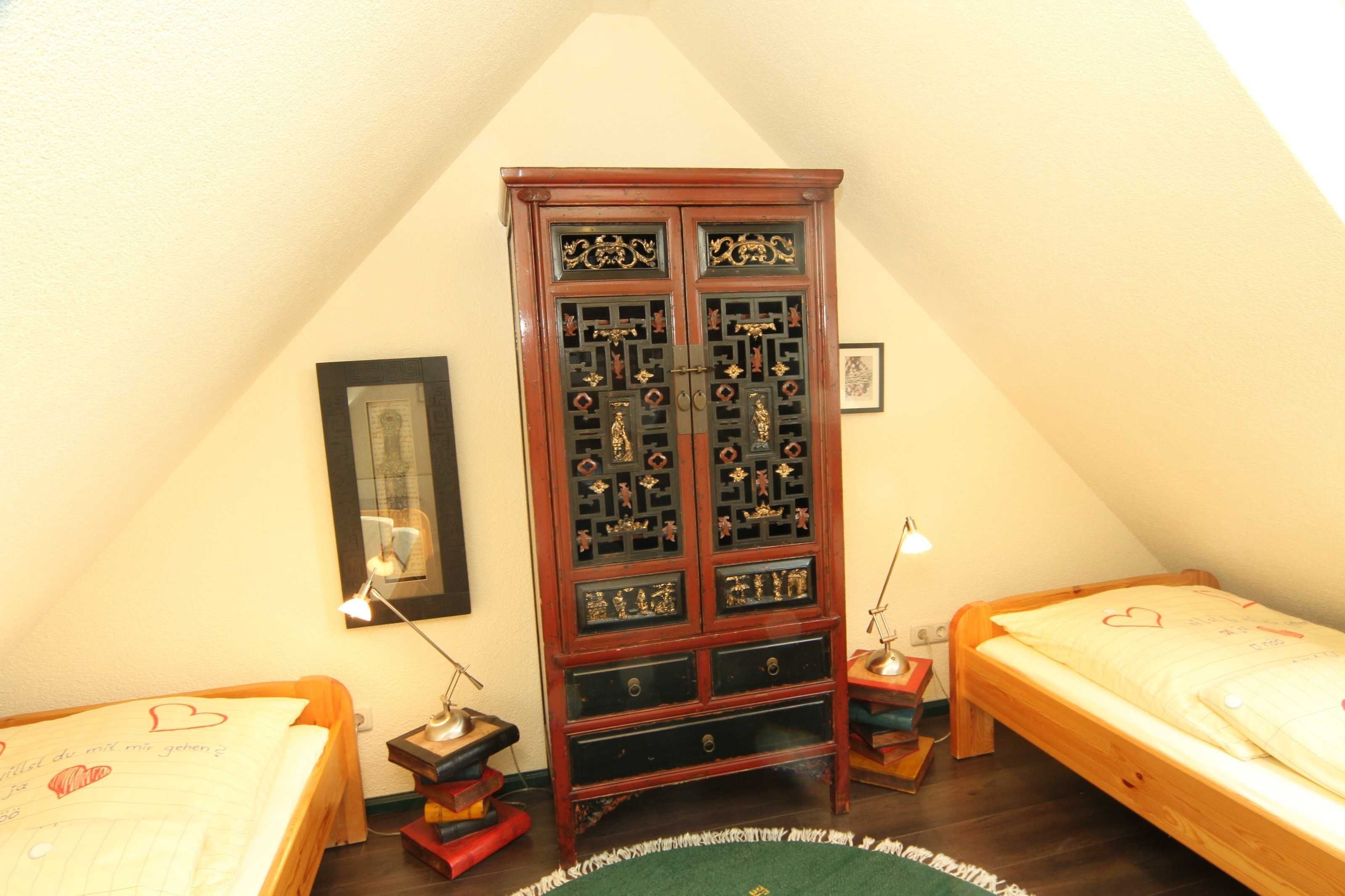 A bedroom with two single beds, a wooden cabinet with intricate designs, a framed picture on the wall, and a small desk with a lamp.