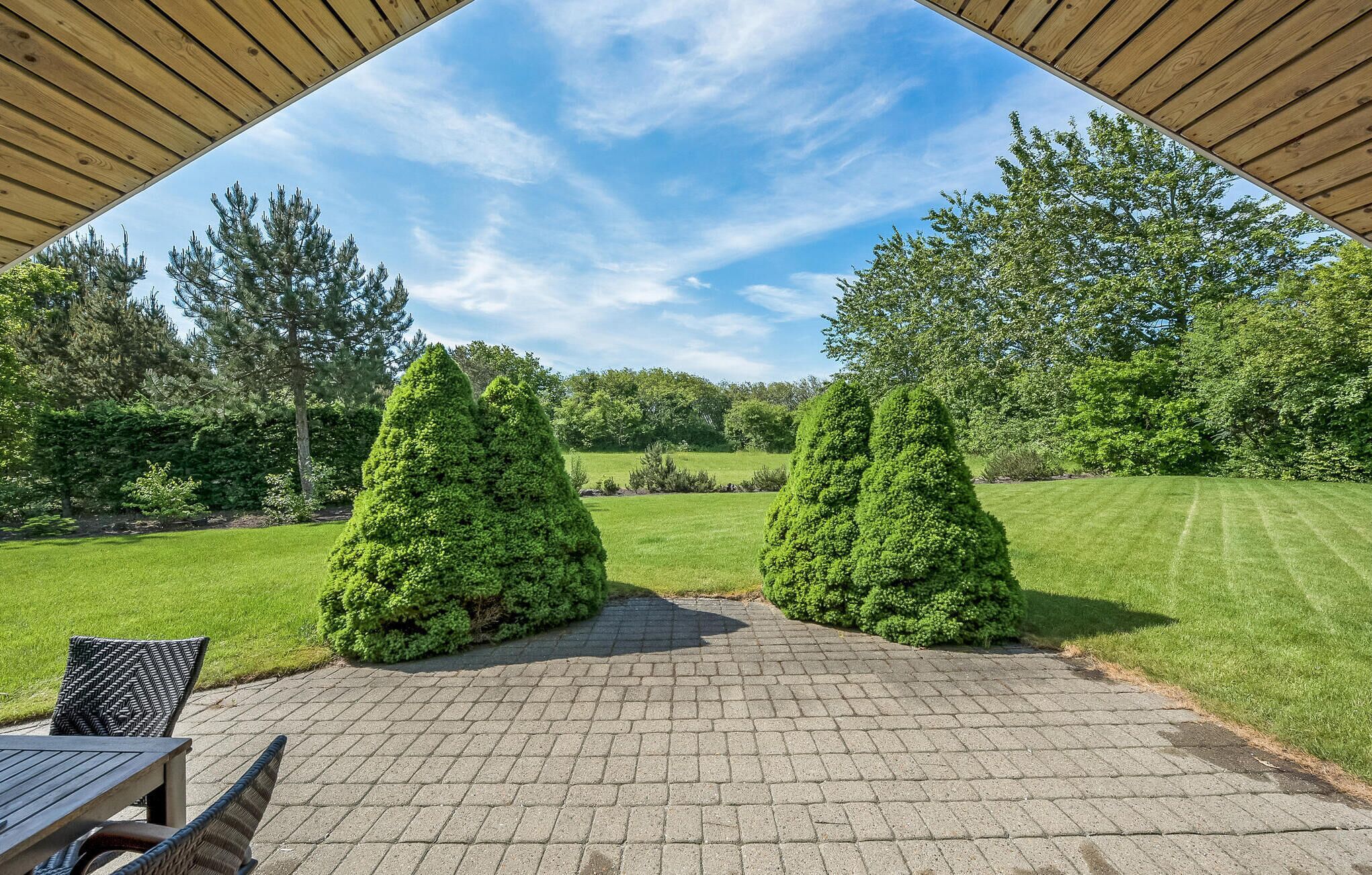 A patio area with a brick floor, two conical topiary trees, a table with chairs, and a view of a grassy field and trees under a clear sky.