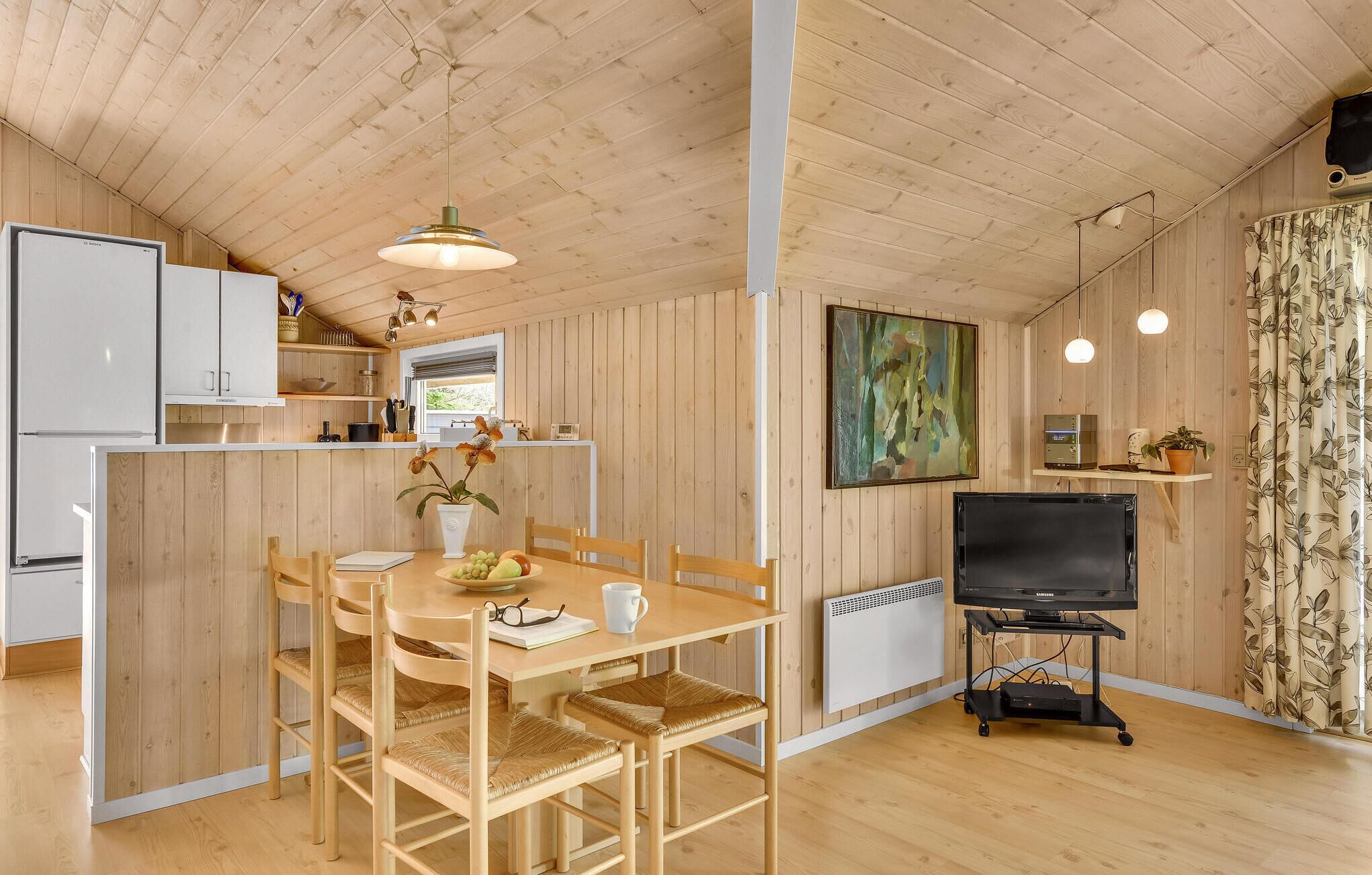 Een compacte keuken met houten kastjes, een eethoek met een tafel en stoelen, een tv op een trolley, en een raam met uitzicht.