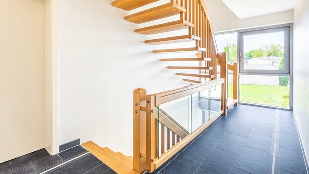 Una escalera moderna con escalones de madera y barandillas de vidrio, que conduce a una habitación con un amplio ventanal con vistas a zonas verdes.