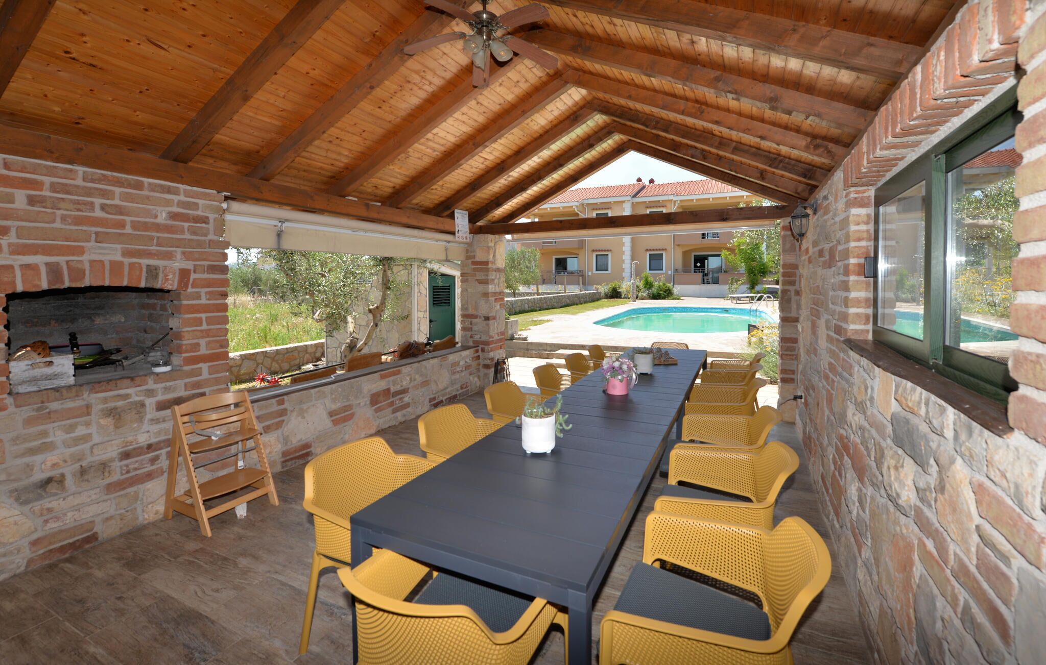 Un patio al aire libre con chimenea de ladrillo, sillas de mimbre amarillo, una mesa larga y vista a una piscina.
