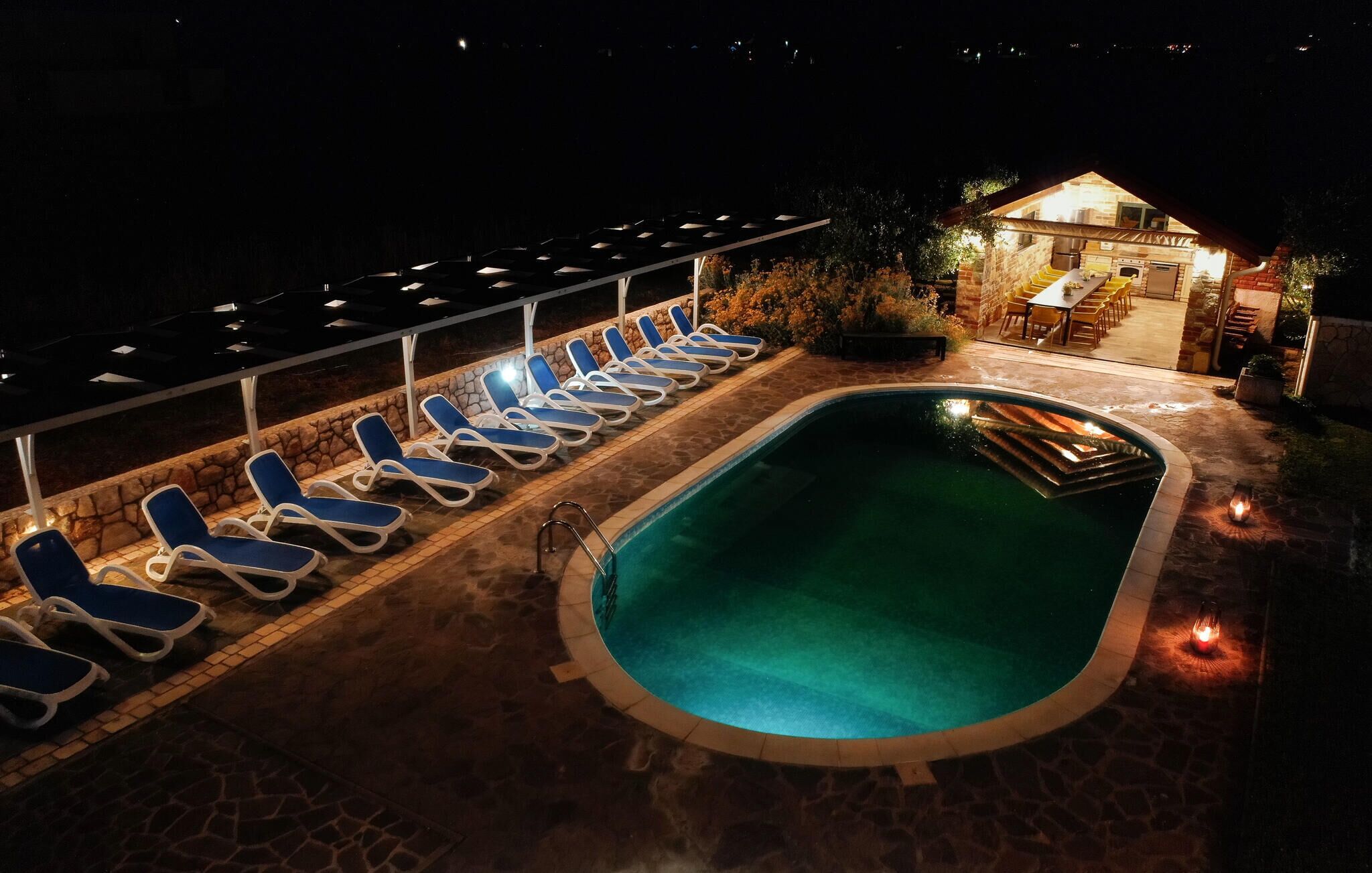 Zona de piscina nocturna con sillones reclinables y un edificio de piedra con asientos al aire libre.