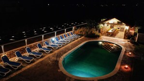 Zona de piscina nocturna con sillones reclinables y un edificio de piedra con asientos al aire libre.