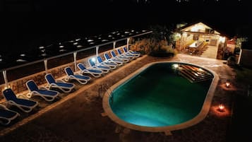Zona de piscina nocturna con sillones reclinables y un edificio de piedra con asientos al aire libre.