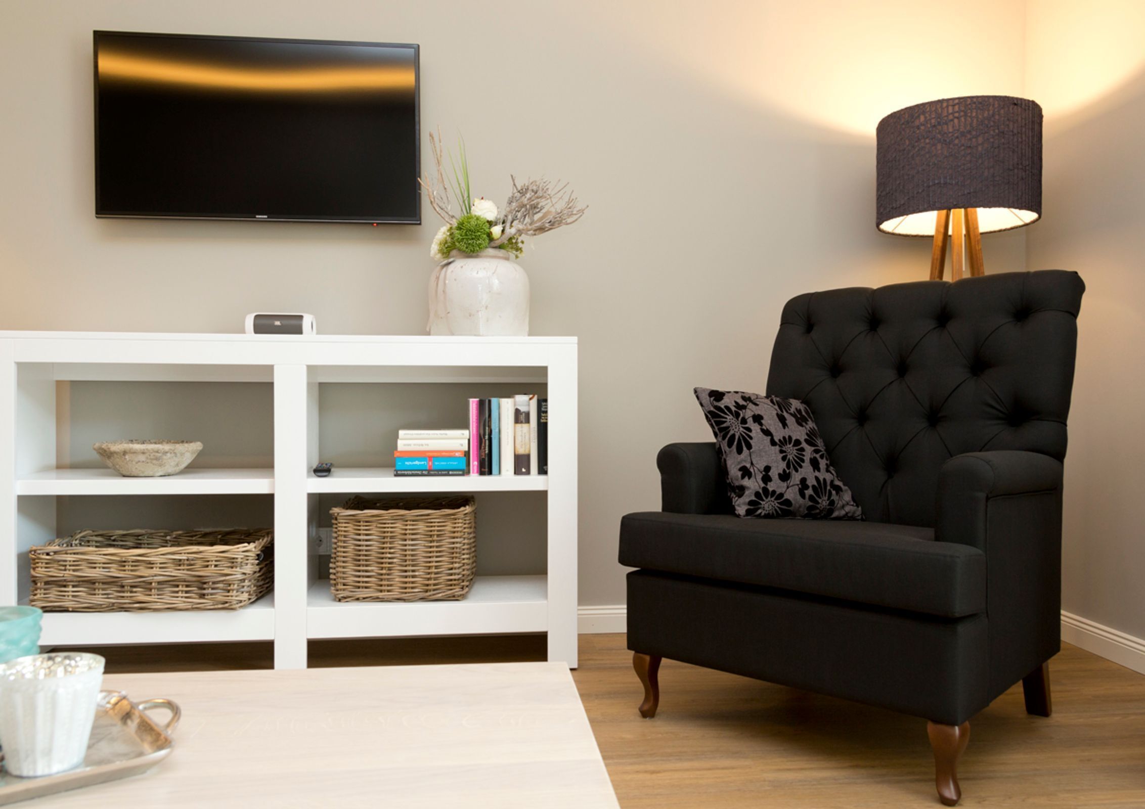 Ett vardagsrum med en svart fåtölj, en vit TV-möbel, en TV, en lampa och en vas med blommor.