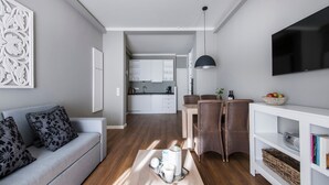 Una sala de estar moderna con un sofá gris, un comedor con sillas de mimbre, una cocina al fondo y un televisor de pantalla plana montado en la pared.