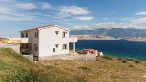 A two-story white building with a red-tiled roof, situated on a grassy hill overlooking a body of water and mountains. - Amazing apartment in Pag with house sea view (Pag)