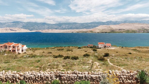 Un paysage côtier avec un mur de pierre au premier plan, une étendue d’eau et des montagnes à l’horizon.