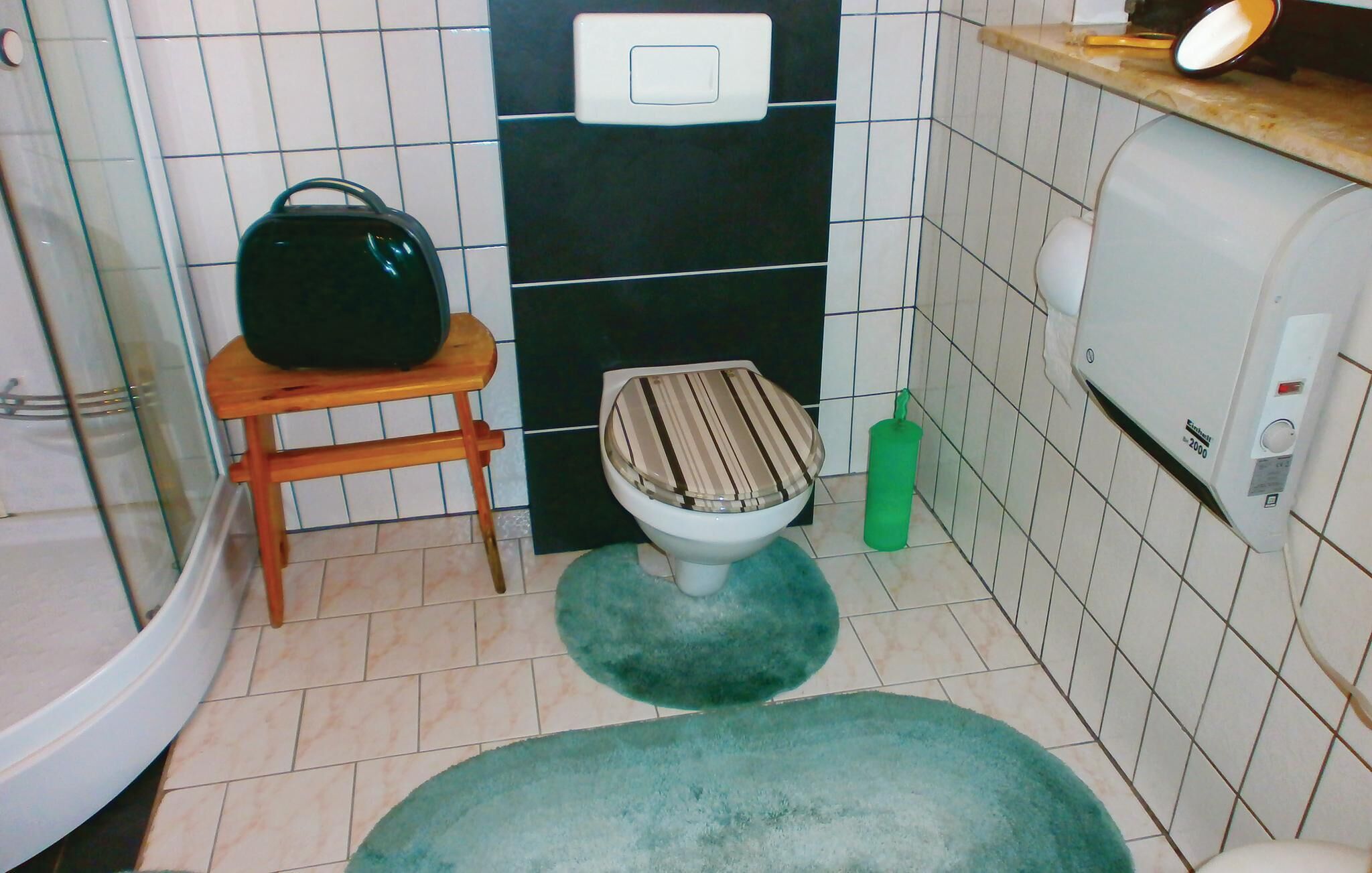 Een kleine badkamer met een toilet, een houten krukje, een groene kleed en een handdroger, plus een zeepdispenser.