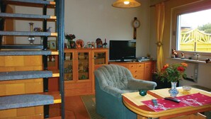 A living room with a blue armchair, a wooden cabinet with glass doors, a television, and a window with curtains.
