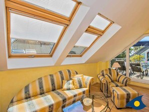 Ein Zimmer mit schrägem Dach und Dachfenstern, ausgestattet mit einer Sofagarnitur und einem Glastisch.