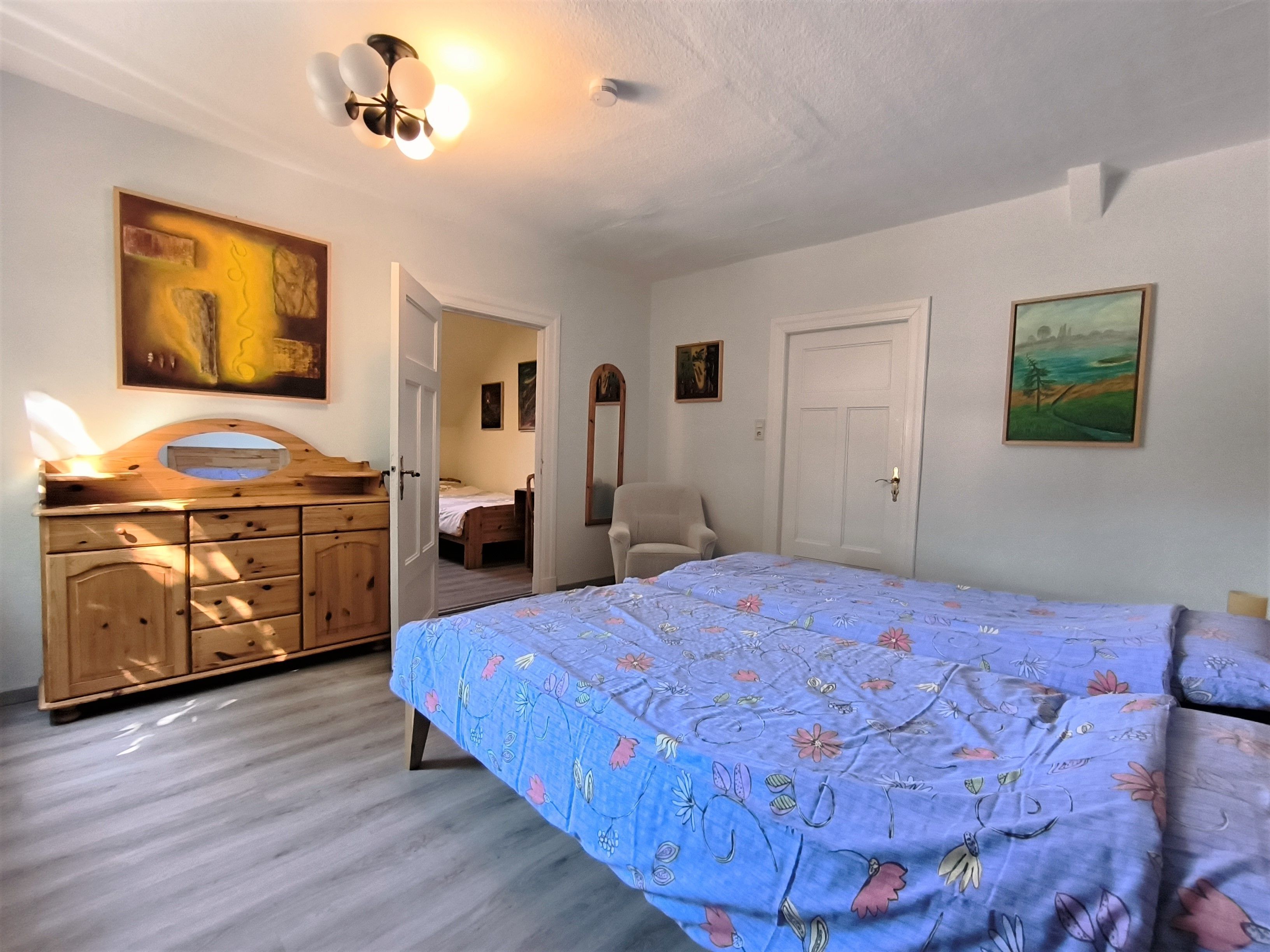 A bedroom with a bed, dresser, chair, and paintings on the walls.