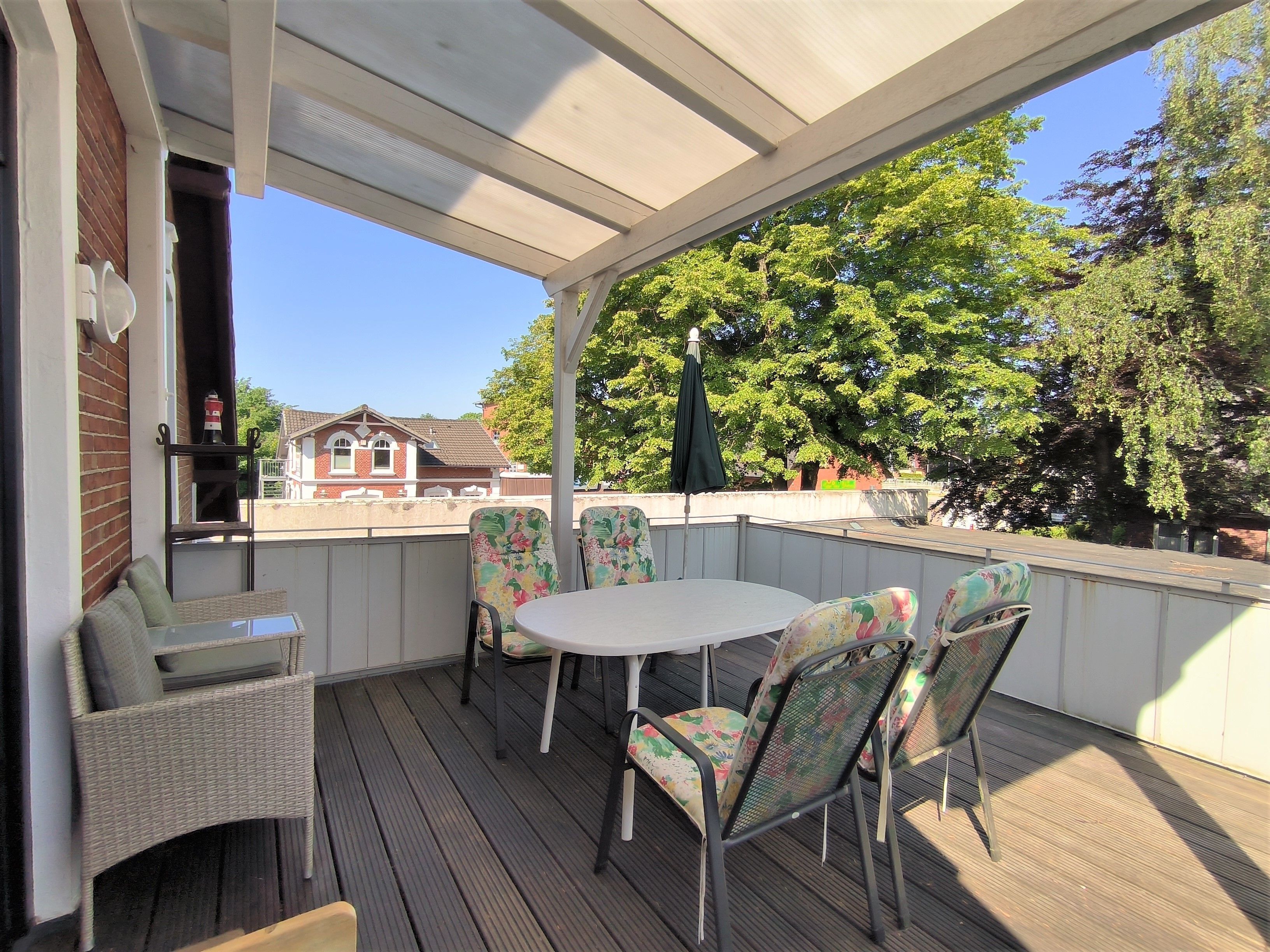 Eine überdachte Terrasse im Freien mit einem runden Tisch und Stühlen, einer Rattan-Couch und Blick auf ein Haus im Hintergrund.