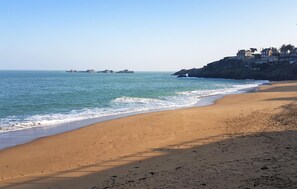 Une plage de sable où l’on peut voir des empreintes de pas menant à l’eau, une falaise rocheuse avec des bâtiments, et une mer calme avec des îles au loin.