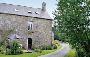 Un bâtiment en pierre avec un toit en pente, un petit porche et un jardin agrémenté de fleurs et d’arbustes.