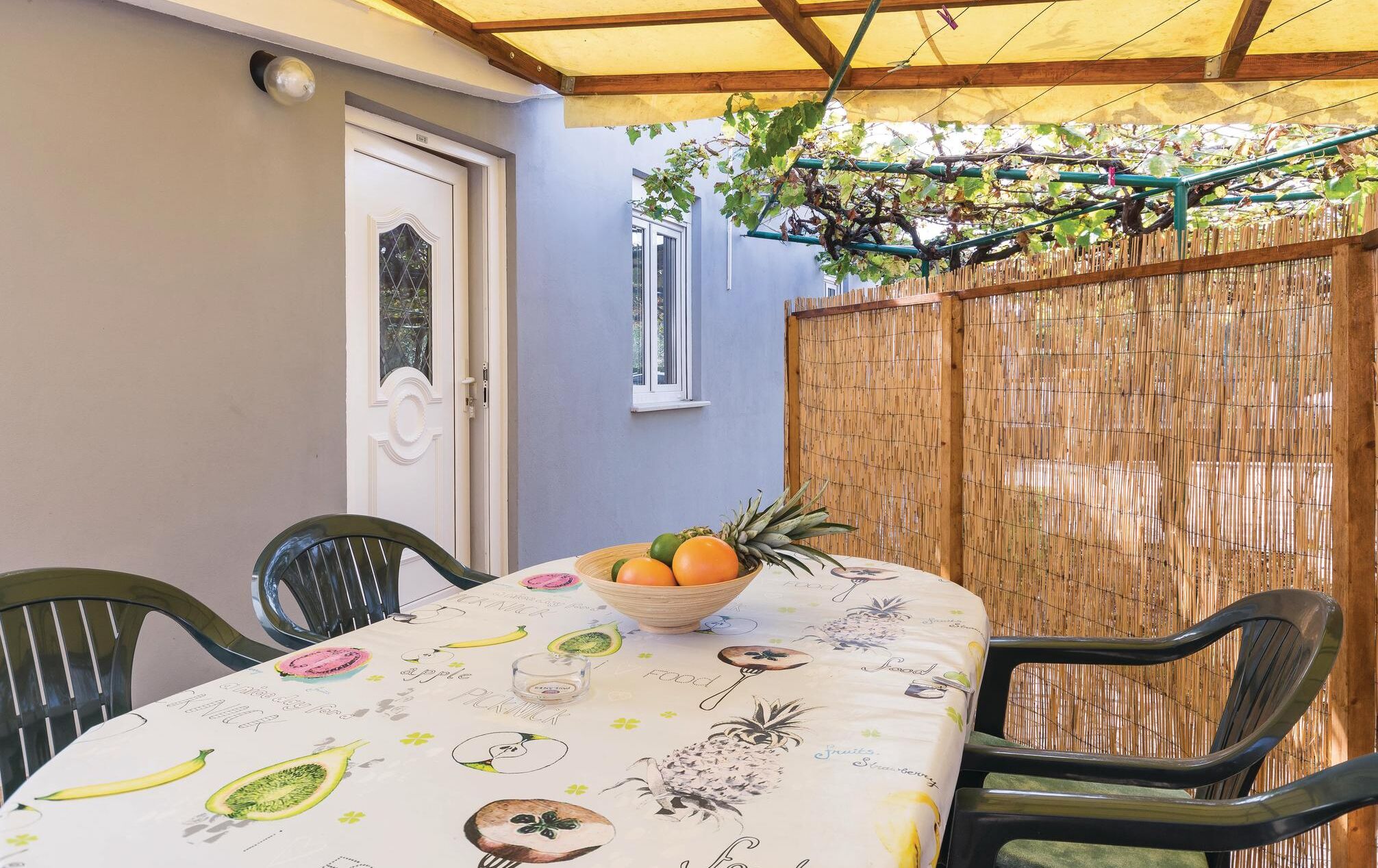 Una mesa con mantel a tema de frutas, un bol de fruta y sillas en un área exterior techada con valla de bambú y una puerta.