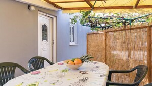 Una mesa con mantel a tema de frutas, un bol de fruta y sillas en un área exterior techada con valla de bambú y una puerta.