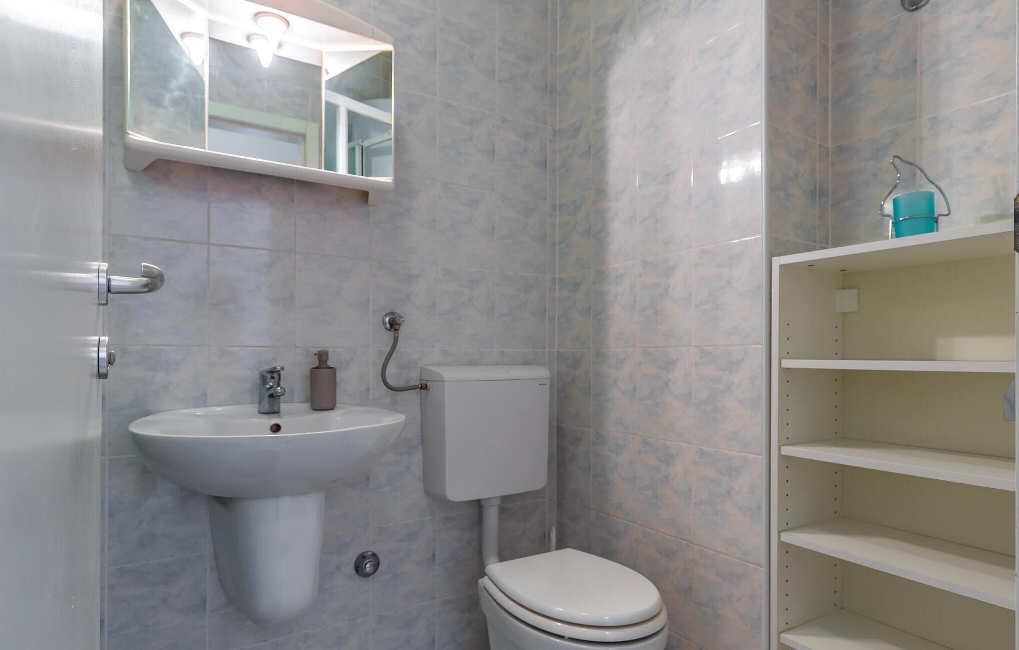 A bathroom with a white sink, a toilet, and a mirror. There are shelves above the toilet.