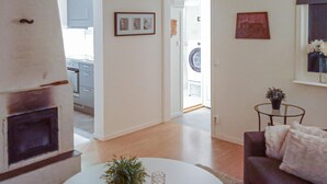 Un salon avec une cheminée, un canapé, une table basse agrémentée d’une plante, et une petite table sur laquelle est posé un vase. Au fond, on aperçoit une cuisine et une porte qui donne sur une autre pièce.